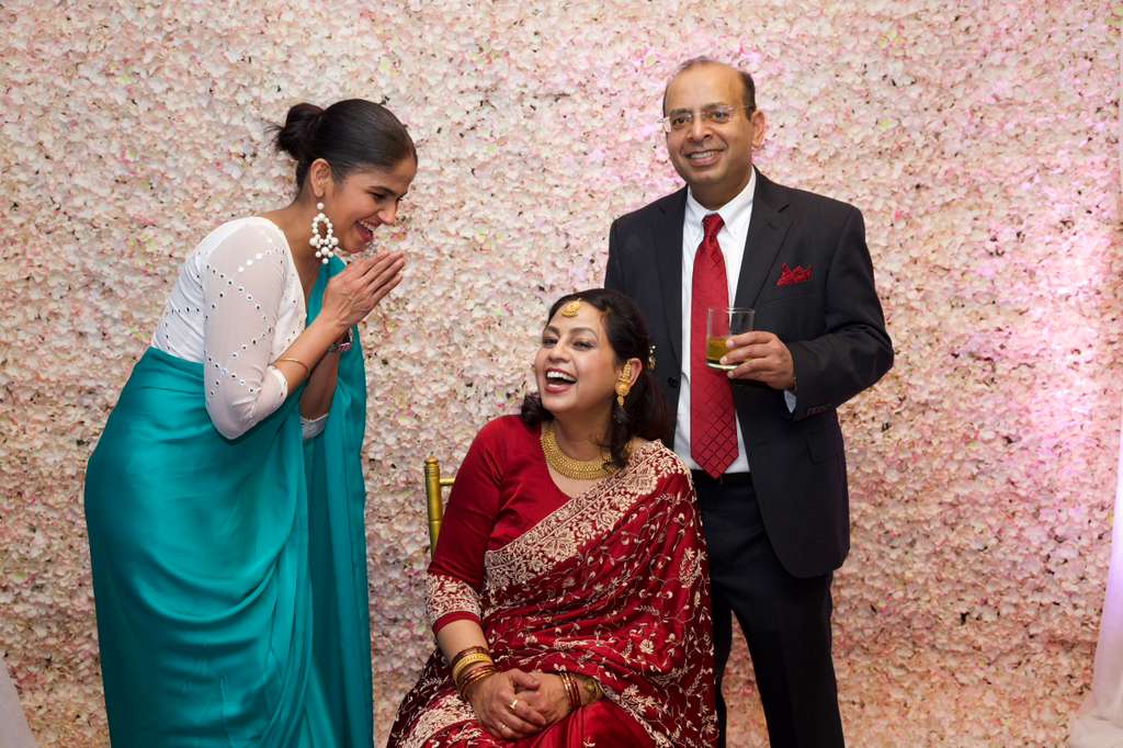 Toolieka in a teal saree sharing a laugh with guests at a wedding reception, floral backdrop